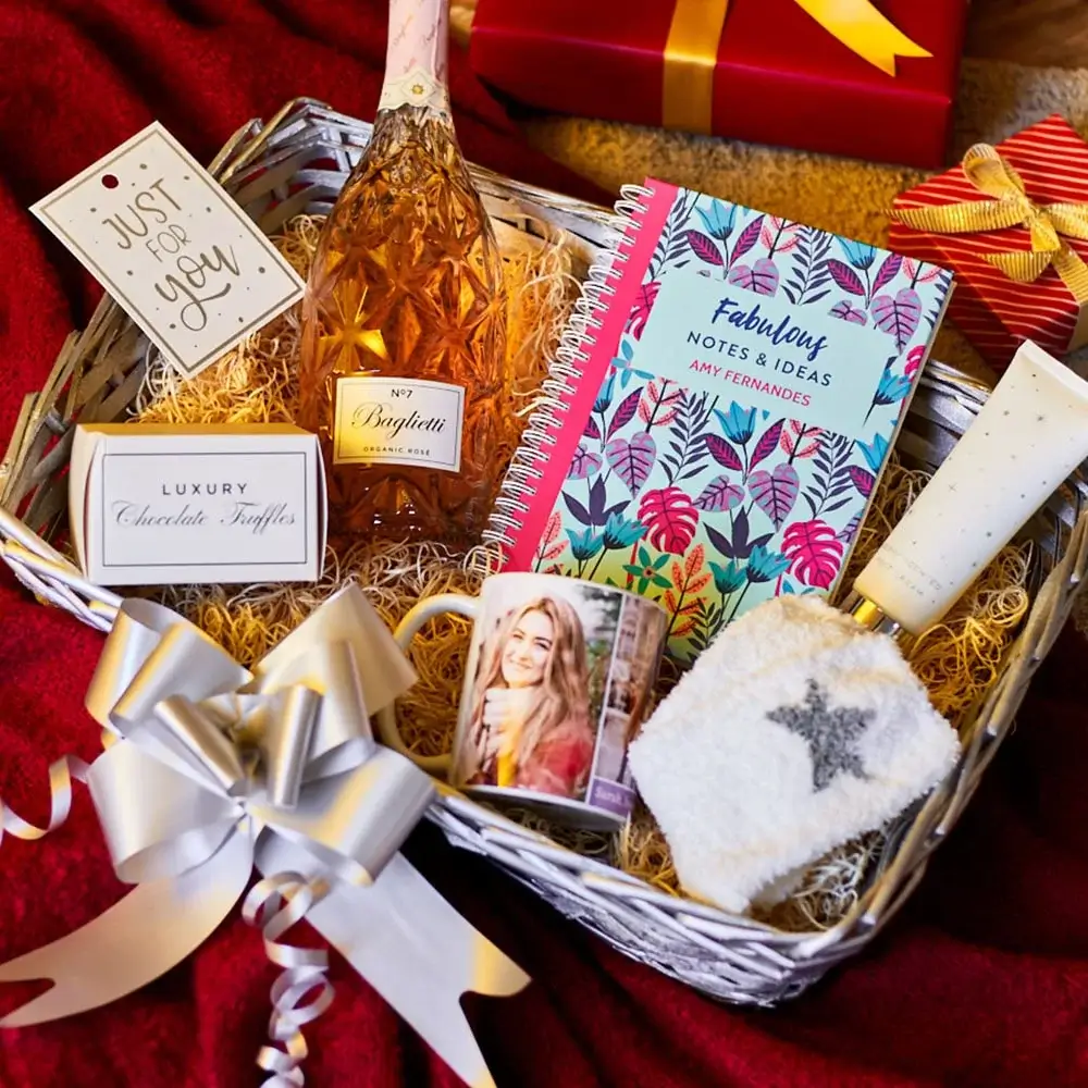 Gift hamper containing a pink sparkling drink, a colorful notebook, luxury chocolate truffles, a mug, cozy socks, a tube of cream, and a decorative card, all arranged in a basket with ribbon and festive packaging in the background.